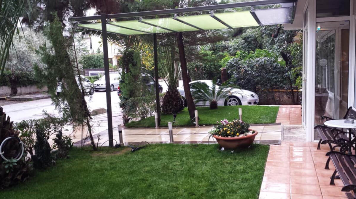 Αυλή της μονάδας με καλυμμένο διάδρομο, φοίνικες, γλάστρες με λουλούδια και σταθμευμένα αυτοκίνητα σε βροχερή μέρα