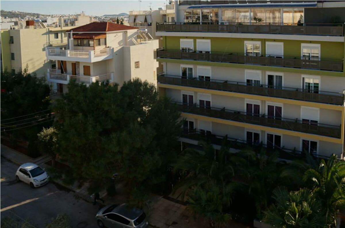 Άποψη της πρόσοψης του κτιρίου με μπαλκόνια, δέντρα και σταθμευμένα αυτοκίνητα στον δρόμο